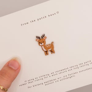 Könnte beinhalten: Ein kleiner gestickter Reh-Aufnäher mit braunem Fell, weißen Punkten und dunkelbraunem Geweih. Der Aufnäher ist auf einer weißen Karte befestigt, die den Text from the patch haus sowie Anweisungen auf Englisch und Deutsch enthält.