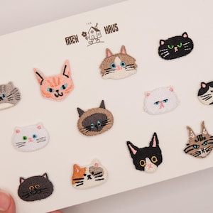 May include: A collection of twelve embroidered cat face patches arranged on a white display card labelled THE PATCH HAUS. The patches feature various feline breeds and colours, including tabby, calico, black, white, and Siamese designs with detailed stitching.