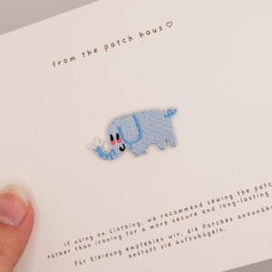 May include: A light blue embroidered elephant patch with pink cheeks and a white flower held in its trunk. The patch is attached to a white card featuring the text from the patch haus and instructions for sewing the patch onto clothing.