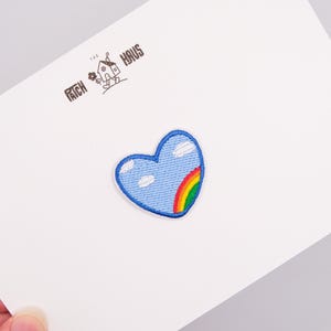 Könnte beinhalten: Ein herzförmiger Stickaufnäher mit hellblauem Himmelshintergrund, weißen Wolken und einem teilweisen Regenbogen auf der rechten Seite. Der Aufnäher ist auf einer weißen Karte befestigt, die mit dem Text THE PATCH HAUS und einem kleinen Haussymbol bedruckt ist.