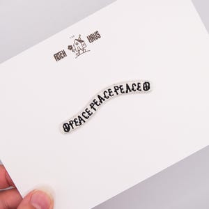 Könnte beinhalten: Ein geschwungener gestickter Aufnäher mit dem dreimal wiederholten Wort PEACE in schwarzem Garn auf weißem Grund. Der Aufnäher ist auf einer weißen Karte befestigt, die mit dem Text THE PATCH HAUS und einer kleinen Hausgrafik bedruckt ist.