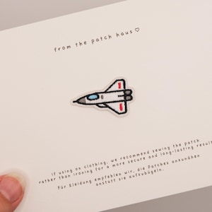 Könnte beinhalten: Ein gestickter Aufbügler in Form eines weißen, grauen und roten Kampfjets mit blauem Cockpitfenster. Der Aufnäher ist auf einer weißen Karte zentriert, die den Text from the patch haus sowie Anweisungen zum Annähen oder Aufbügeln enthält.