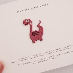 Può includere: Una piccola toppa ricamata a forma di dinosauro nei toni del rosa scuro e bordeaux con un bordo bianco. La toppa è fissata su un cartoncino bianco con il testo from the patch haus e le istruzioni per cucire la toppa sui capi di abbigliamento.