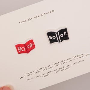 May include: Two embroidered patches on a white card. One patch is red with white text reading "Bo ok" shaped like an open book. The other patch is black with white text reading "Bo || ok" shaped like an open book. The card includes the text "from the patch haus" and instructions for application.