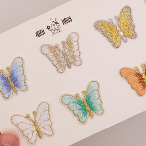 Op de afbeelding: Zes geborduurde vlinderpatches gerangschikt op een witte kaart. De vlinders hebben verschillende kleuren, waaronder blauw, wit, goud, turquoise en oranje met details van metaaldraad. De tekst THE PATCH HAUS staat in het zwart bovenaan gedrukt.