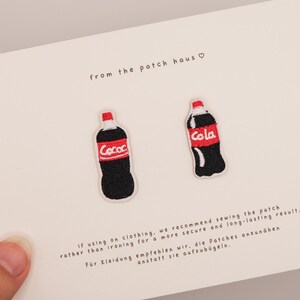 Pode incluir: Dois emblemas bordados em forma de garrafas de refrigerante pretas com rótulos vermelhos e texto branco. Os emblemas estão fixados num cartão branco com o texto from the patch haus, juntamente com instruções em inglês e alemão sobre a aplicação.