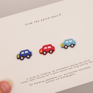 Könnte beinhalten: Drei kleine gestickte Auto-Aufnäher in Dunkelblau, Rot und Hellblau auf einer weißen Karte mit dem Text from the patch haus und Anwendungshinweisen. Jedes Auto hat schwarze Räder und gelbe Scheinwerfer als gestickte Details.