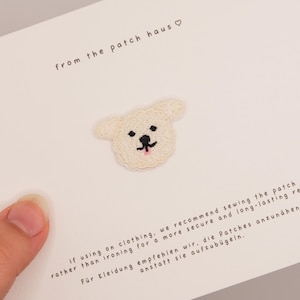 Puede incluir: Un pequeño parche bordado de color crema con forma de cabeza de perro, con ojos negros, nariz negra y una diminuta lengua rosa. El parche está centrado en una tarjeta blanca con el texto from the patch haus e instrucciones de costura.