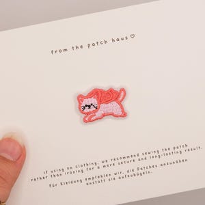 Pode incluir: Um emblema bordado de gato com corpo rosa claro e capa vermelho coral, exibido num cartão branco com o texto from the patch haus e instruções que recomendam costurar o emblema para um resultado seguro e duradouro.