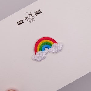 May include: An embroidered patch featuring a colorful rainbow with red, yellow, green, and blue stripes, flanked by two white clouds. The patch is attached to a white card printed with the black text THE PATCH HAUS and a small house icon.