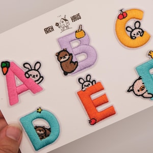 Op de afbeelding: Een set van zes geborduurde alfabet-patches van A tot F op een witte kaart met de tekst The Patch Haus. Elke letter heeft een unieke kleur en een klein geborduurd dier of voedselitem, waaronder een konijn, otter, kers en eendje.