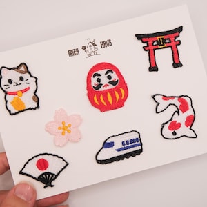 May include: A set of embroidered patches on a white card featuring a beckoning cat, a red Daruma doll, a red torii gate, a pink cherry blossom, a white bullet train, a koi fish, and a folding fan with a red circle. The text Patch Haus is printed.