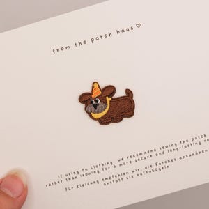 Puede incluir: Un parche bordado de un perro marrón con un gorro de fiesta naranja y un collar amarillo, centrado en una tarjeta blanca. La tarjeta incluye el texto from the patch haus e instrucciones para coser el parche en inglés y alemán.