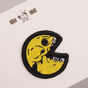 Op de afbeelding: Een ronde geborduurde patch met een gele, schedelachtige figuur met een wijd open mond die witte tanden laat zien, omlijnd met zwart garen. Boven de patch staat de tekst THE PATCH HAUS gedrukt met een klein icoontje van een huisje.