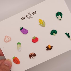 Könnte beinhalten: Eine Sammlung kleiner gestickter Gemüseaufnäher auf einer weißen Karte mit dem Text PATCH HAUS. Das Set enthält eine Karotte, Knoblauch, eine Aubergine, Mais, eine Tomate, Brokkoli, einen Pilz und weitere verschiedene grüne Gemüsemotive.