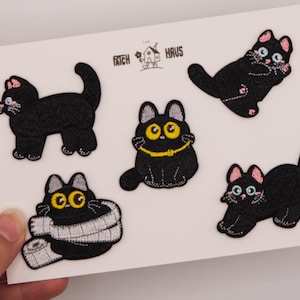 May include: Five embroidered black cat patches on a white backing card with the text THE PATCH HAUS. The cats feature various poses, including one with a yellow collar, one wrapped in a white scarf, and others with pink ears and large eyes.