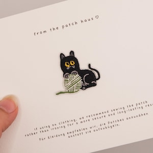 May include: An embroidered patch featuring a black cat with yellow eyes playing with a green and white yarn ball. The patch is attached to a white card with the text from the patch haus and instructions in English and German regarding application.