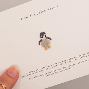 Può includere: Una toppa ricamata a forma di pinguino con testa grigio scuro, faccia bianca, pancia marrone chiaro e piedi gialli. La toppa è centrata su un cartoncino bianco stampato con il testo from the patch haus e le istruzioni per l'applicazione.
