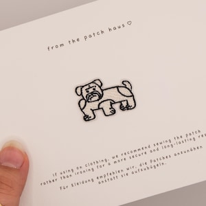 Könnte beinhalten: Ein weißer Stickaufnäher mit der schwarzen Umrisszeichnung einer Bulldogge, zentriert auf einer weißen Karte. Die Karte enthält den Text from the patch haus sowie Anweisungen auf Englisch und Deutsch zum Annähen des Aufnähers auf Kleidung.