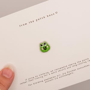 Op de afbeelding: Een kleine, groene, geborduurde kikkergezicht-patch gecentreerd op een witte kaart. De kaart bevat de tekst from the patch haus met een hartsymbool en instructies in het Engels en Duits over het naaien van de patch op kleding.