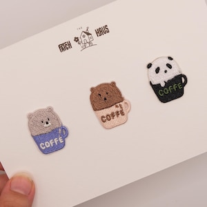 May include: Three embroidered patches featuring animal-themed coffee mugs on a white card with the text THE PATCH HAUS. The patches depict a grey bear in a blue mug, a brown bear in a light pink mug, and a panda in a black mug, all labelled COFFE.