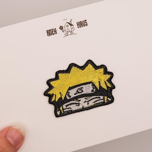 Op de afbeelding: Een geborduurde patch met een gestileerd personage met stekelig geel haar en een grijze hoofdband met een zwart spiraalsymbool. De patch is bevestigd op een witte kaart met de tekst THE PATCH HAUS en een kleine illustratie van een huis.