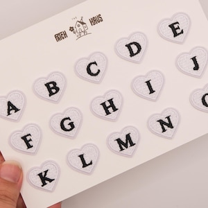 May include: A white card featuring fifteen heart-shaped embroidered patches, each displaying a black letter from A to O. The patches are arranged in rows, and the card includes the text THE PATCH HAUS with a small house illustration at the top.