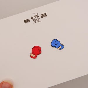 Könnte beinhalten: Zwei kleine gestickte Aufnäher in Form von Boxhandschuhen, einer rot und einer blau, präsentiert auf einer weißen Karte mit dem Text THE PATCH HAUS und einer kleinen Hausillustration, die in schwarzer Tinte oben mittig aufgedruckt ist.