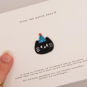 Op de afbeelding: Een kleine geborduurde patch in de vorm van een zwarte kattenkop met een blauw en groen gestreept feesthoedje met een rode pompon. De patch is bevestigd op een witte kaart met de tekst from the patch haus en instructies voor het naaien.