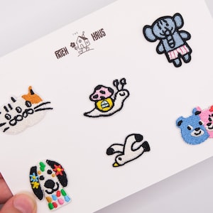 Op de afbeelding: Een verzameling van zes geborduurde patches met een kattengezicht, een slak met een paddenstoelhuisje, een blauwe olifant, een meeuw, een hond met kleurrijke vlekken en twee berengezichten. De tekst THE PATCH HAUS staat op de witte kaart.