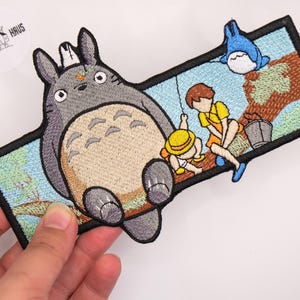 Könnte beinhalten: Ein rechteckiger bestickter Aufnäher mit einer großen grauen Kreatur mit beigem Bauch, zwei Kindern in gelber und orangefarbener Kleidung sowie einer kleinen blauen Kreatur. Das Motiv zeigt einen braunen Ast, einen grauen Eimer und hellblauen Himmel.