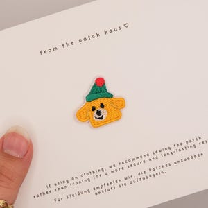 Könnte beinhalten: Ein gestickter Aufnäher mit einem gelben Hund, der eine grüne Mütze mit rotem Bommel trägt. Der Aufnäher ist auf einer weißen Karte befestigt, die mit dem Text from the patch haus und Anweisungen zum Annähen auf Kleidung bedruckt ist.