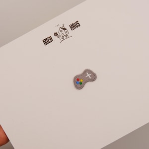 Pode incluir: Um pequeno emblema bordado cinza em forma de controle de videogame com uma cruz direcional branca e quatro botões coloridos em vermelho, azul, amarelo e verde. O emblema está preso a um cartão branco com o texto THE PATCH HAUS.