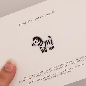 May include: A small embroidered zebra patch featuring black and white stripes and a tiny pink cheek detail. The patch is attached to a white display card with the text from the patch haus and instructions for sewing the patch onto clothing.