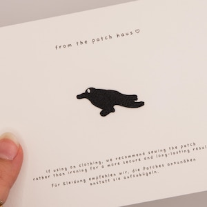Puede incluir: Un parche bordado negro con forma de pájaro centrado en una tarjeta blanca. La tarjeta incluye el texto from the patch haus con un símbolo de corazón e instrucciones en inglés y alemán sobre cómo coser el parche en la ropa.