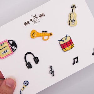 Op de afbeelding: Een verzameling geborduurde patches met muziekthema's, waaronder een gele gitaar, een gele trompet, een rood-gele trommel, een zwarte koptelefoon, een microfoon en muzieknoten. De tekst THE PATCH HAUS staat erboven gedrukt.