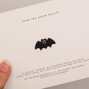 May include: A small black embroidered bat patch centered on a white card. The card features the text from the patch haus and instructions in English and German regarding sewing the patch onto clothing for a secure and long-lasting result.