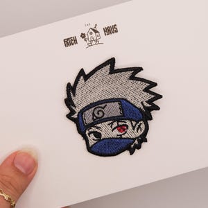 Könnte beinhalten: Ein gestickter Aufnäher mit einem stilisierten Anime-Charakter mit stacheligem silbernem Haar, einem blauen Stirnband mit Spiralsymbol, einer blauen Gesichtsmaske und einem roten Auge. Der Aufnäher ist auf einer weißen Karte mit der Aufschrift THE PATCH HAUS befestigt.