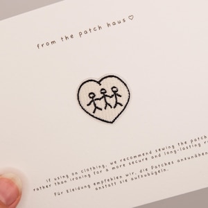 May include: A white heart-shaped embroidered patch featuring three black stick figures holding hands. The patch is attached to a white card with the text from the patch haus and instructions for sewing the patch onto clothing.
