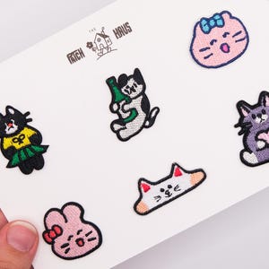 Puede incluir: Un conjunto de seis parches bordados sobre una tarjeta blanca con el texto THE PATCH HAUS. Los diseños incluyen un gato negro con camisa amarilla, un gato sosteniendo una botella verde, una cara de gato rosa, un gato morado con un corazón, una cabeza de gato blanca y un conejo rosa.
