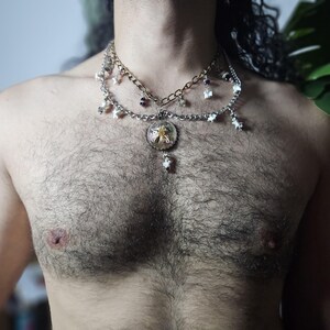 Könnte beinhalten: Eine mehrlagige Halskette mit einer goldfarbenen und einer silberfarbenen Kette. Die silberne Kette ist mit kleinen weißen sternförmigen Anhängern und einem runden Mittelanhänger verziert, der eine gelbe Blume in klarem Harz enthält.