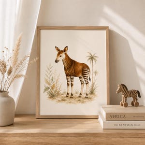 Op de afbeelding: Een ingelijste kunstdruk met een aquarelillustratie van een okapi die in een grasachtig landschap met palmbomen staat. De okapi heeft een bruin lichaam met wit en zwart gestreepte poten. Het kunstwerk zit in een lichthouten lijst.