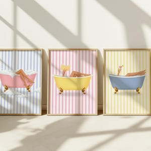 Puede incluir: Un conjunto de tres láminas decorativas enmarcadas con bañeras de patas. La primera muestra una bañera rosa sobre fondo de rayas azules con espuma, la segunda una bañera amarilla sobre rayas rosas con un libro, y la tercera una bañera azul sobre rayas amarillas con una bebida.