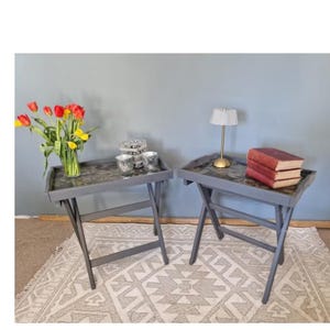 May include: Two grey folding tray tables with dark, mottled surfaces. The left table holds a clear glass vase with red and yellow tulips and two small glass votives. The right table holds a small gold-toned lamp and three stacked books.