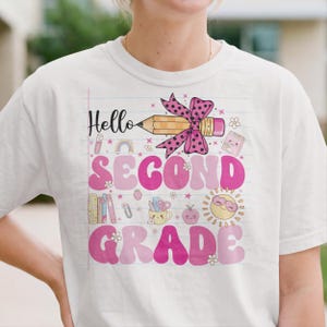 May include: A white t-shirt featuring a graphic print on a lined paper background. The design includes the text "Hello SECOND GRADE" in pink, a yellow pencil with a pink polka-dot bow, and small illustrations of books, a sun, and school supplies.