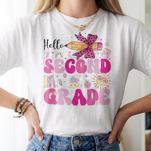 May include: A white t-shirt featuring a graphic print on a lined paper background. The design includes a pencil with a pink polka dot bow, the text Hello SECOND GRADE in pink, and various icons like books, a sun, and a mug with school supplies.