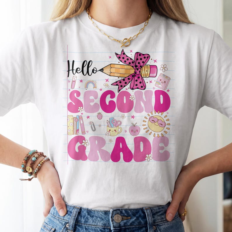 May include: A white t-shirt featuring a graphic print on a lined paper background. The design includes a pencil with a pink polka dot bow, the text Hello SECOND GRADE in pink, and various icons like books, a sun, and a mug with school supplies.