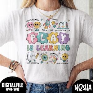 May include: A white t-shirt featuring a colorful graphic with the text PLAY IS LEARNING. The design includes cartoon illustrations of a rocket, a bead maze, a notebook, a magnifying glass, a paint palette, and a lightbulb with labels.