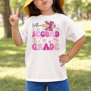 May include: A white short-sleeve t-shirt featuring a graphic print with the text Hello SECOND GRADE. The design includes a yellow pencil with a pink polka-dot bow, a sun, books, and various school-themed icons in shades of pink and yellow.