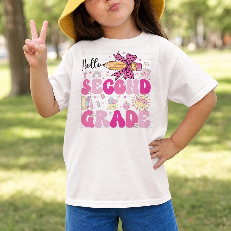 May include: A white short-sleeve t-shirt featuring a graphic print with the text Hello SECOND GRADE. The design includes a yellow pencil with a pink polka-dot bow, a sun, books, and various school-themed icons in shades of pink and yellow.